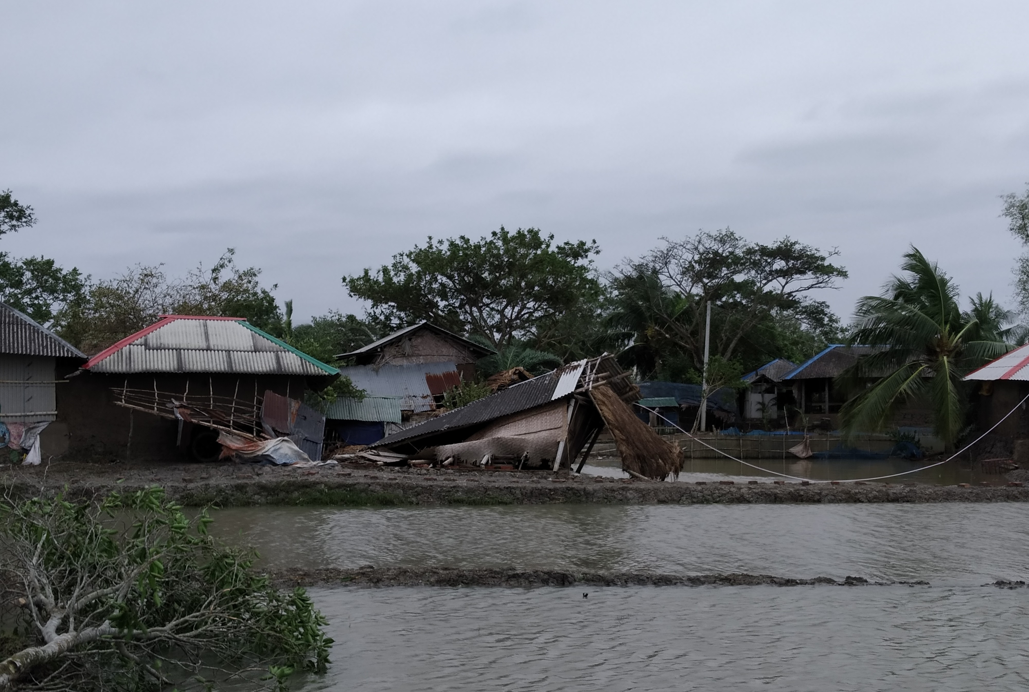 A photo of a row of small houses stands beside a flooded area, with one structure partially collapsed and leaning into the water. Debris is scattered around the damaged building. Trees and other intact houses surround the scene under an overcast sky.